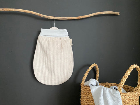 Ein KraftKids Strampelsack Frühling Sommer Cord Breitcord in Beige Taupe hängt an einem Holzast vor einer dunklen Wand. Ein geflochtener Korb mit Griffen darunter hält ein hellgraues Tuch.