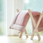 Eine KraftKids Krabbeldecke Musselin in zartem Rosa ist über einen hellen Holzständer an einem großen Fenster drapiert, daneben steht ein mit rosa Bommeln verzierter Strohkorb auf dem hellen Holzboden.