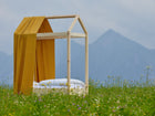 Ein minimalistisches Holzbettgestell mit dem KraftKids Bettbaldachin Musselin gelb Pusteblumen und weißem Bettzeug steht in einem Wildblumenfeld, im Hintergrund erheben sich neblige Berge unter einem wolkenverhangenen Himmel.
