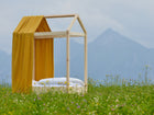 Ein minimalistisches Hausbett mit dem KraftKids Bettbaldachin Musselin goldene Punkte auf Gelb und weißer Bettwäsche steht in einer Wildblumenwiese vor Bergen unter bewölktem Himmel.