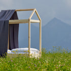 Ein minimalistischer Holzbettrahmen mit dem KraftKids Bettbaldachin Musselin goldene Punkte auf Grau ist elegant drapiert, eingebettet in eine Wildblumenwiese mit Bergen im Hintergrund unter einem wolkenverhangenen Himmel.