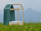 Ein einfaches Holzbett mit weißem Bettzeug und dem Bettbaldachin Musselin nilblau von KraftKids steht auf einer grünen Wiese mit Wildblumen, Bergen und einem wolkenverhangenen Himmel im Hintergrund.