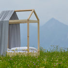 Ein KraftKids Bettbaldachin Musselin grau Pusteblumen, gepaart mit weißer Bettwäsche, steht in einer Wildblumenwiese vor Bergen und Wolken - eine bezaubernde, von der Natur inspirierte Dekoration für das Kinderzimmer.