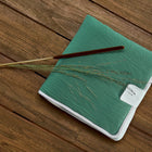 Eine grün strukturierte KraftKids Reisewickelunterlage Doppelkrepp Jade mit weißem Etikett sitzt auf Holz, gekrönt von einem braunen Stock und getrocknetem Gras - ideal, um Notizen zu machen, während Sie Ihre Wickelunterlage einpacken.