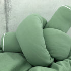 Eine Nahaufnahme des KraftKids Wickeltischumrandung Doppelkrepp Grün Jade, ein weiches grünes geflochtenes Kissen mit weißer Paspel, zu einem Knoten gedreht und vor einem hellgrauen Hintergrund.