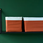 Zwei KraftKids Körbchen Doppelkrepp Rot Herbstrot mit orangefarbenen Böden und weißen Oberteilen sitzen nebeneinander auf einem schwarzen Regal vor einer dunkelgrünen Wand - ideal zur Aufbewahrung auf einem Wickeltisch.