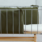 Eine Nahaufnahme eines modernen Babybettchens mit grauem Metallrahmen, KraftKids Bettumrandung Nestchen Doppelkrepp Grün Herbstgrün an den Stäben und einer gefalteten weißen Decke auf einem hellen Holzsockel.