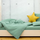 Ein helles Holzbett mit KraftKids Bettwäscheset Doppelkrepp Grün Jade aus 100% Baumwolle und einem gelben Sternenkissen steht auf einem Holzboden an einer Betonwand. Die handgefertigte Bettwäsche verspricht einen erholsamen Schlaf.