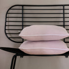 Zwei Kissenbezüge Doppelkrepp Rosa von KraftKids mit weißem Rand sind auf einem modernen schwarzen Metallstuhl mit Gitterrückenlehne vor einer neutralen beigen Wand gestapelt.