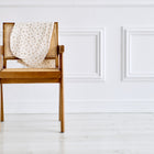 Vor einer weiß getäfelten Wand steht ein Holzstuhl mit Schilfrohr-Details, der mit einem KraftKids Stillkissenbezug Musselin Zweige braun mit zartem Zweigmuster drapiert ist. Der helle Holzboden trägt zu der beruhigenden Atmosphäre bei.