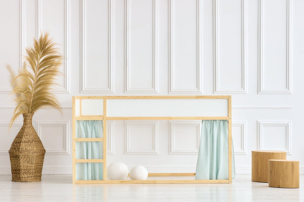 Ein minimalistisches Kinderbett mit KraftKids Hochbettvorhänge Set 2+1 Musselin mint Pusteblumen, kompatibel mit dem Kura Bett, steht in einem hellen weißen Raum mit getäfelten Wänden. In der Nähe befinden sich eine Weidenvase mit Pampasgras, zwei Holzhocker und zwei weiße Bälle unter dem Bett.
